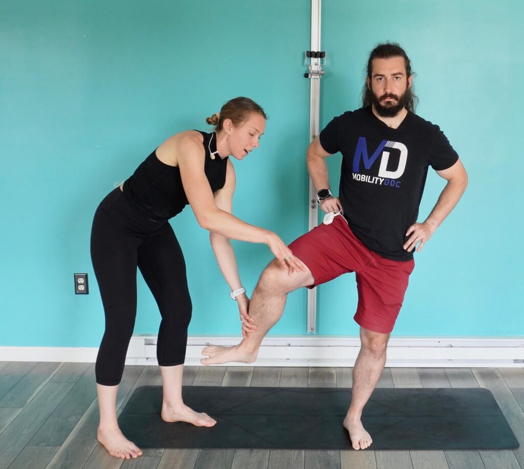 Dr. John demonstrating hip circles to activate and open his glutes. Dr. Chloe is taking his free leg at the knee and foot and is helping to demonstrate the proper movement to turn his hips